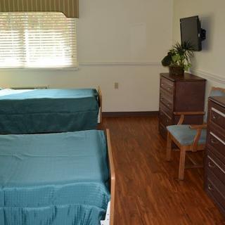 A hospital room with two beds, a wall-mounted television, a dresser, and a window with curtains.