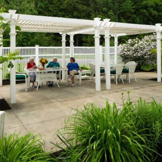 A serene garden patio with a white pergola and outdoor seating, surrounded by lush greenery.