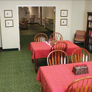 A cozy indoor dining area with tables covered in red tablecloths, wooden chairs, and a bookshelf against a carpeted floor.