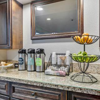 A well-organized coffee station with beverage dispensers, a fruit basket, and cups on a countertop.