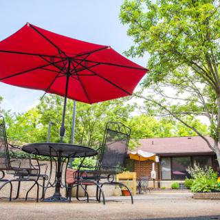 A bright red umbrella provides shade over a patio table and chairs, surrounded by greenery.