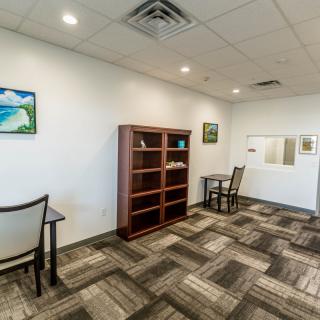 A tidy office space with a bookshelf, desks, and chairs, adorned with paintings on the walls and carpeted flooring.
