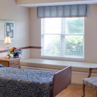 A cozy bedroom featuring a single bed, bedside table with a lamp, and a window seat providing natural light.