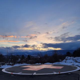 A scenic view of a helipad at sunset, surrounded by snow-covered mountains and dramatic clouds.