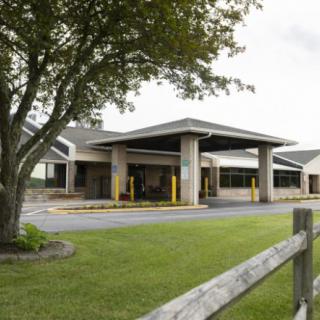 A modern building with a covered entrance, surrounded by a landscaped area with trees and a wooden fence.