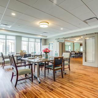A modern dining room with wooden flooring, featuring a set of tables and chairs, illuminated by ceiling lights and surrounded by large windows.