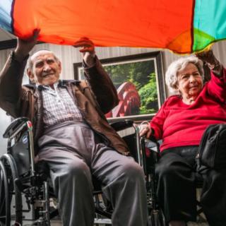 Elderly couple participating in a colorful parachute activity indoors.