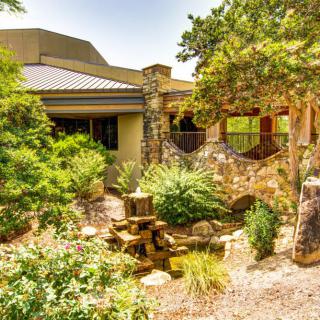 A tranquil garden landscape featuring lush greenery, a stone building, and a small water feature.