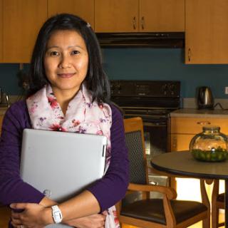 A woman holding a laptop, smiling in a cozy kitchen setting.