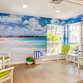 A bright room with a beach-themed mural, featuring chairs, a table, and windows letting in natural light.