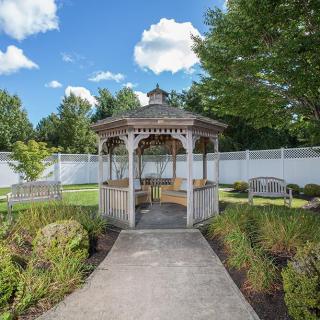 A scenic garden view featuring a wooden gazebo, surrounded by lush greenery and benches under a clear blue sky.