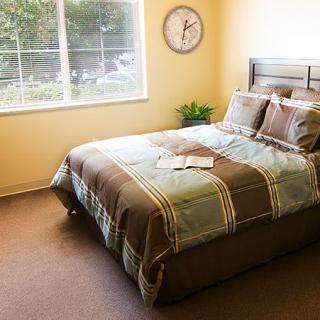 A cozy bedroom with a neatly made bed, a wall clock, and a window letting in natural light.