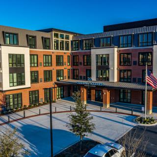 A modern apartment complex with large windows and an entrance area, featuring a flagpole and landscaped surroundings.