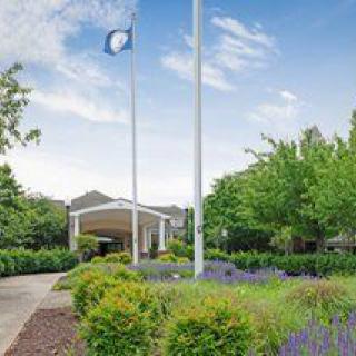 A building entrance surrounded by lush greenery and a flagpole, under a clear blue sky.