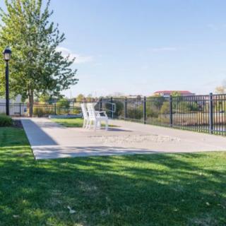A peaceful outdoor patio area with white chairs, surrounded by a grassy park and trees under a clear blue sky.
