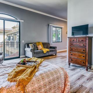 A cozy bedroom with modern decor, featuring a bed with a patterned blanket, a wooden dresser, and ample natural light from large windows.