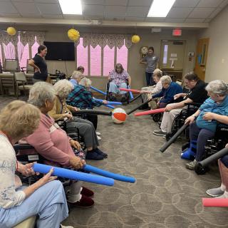 A group of seniors participating in a fun indoor exercise activity involving bright foam sticks and a large ball.