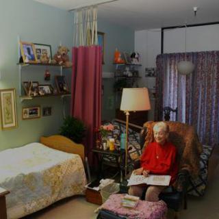 An elderly woman sitting in a cozy room decorated with various personal items and furniture.