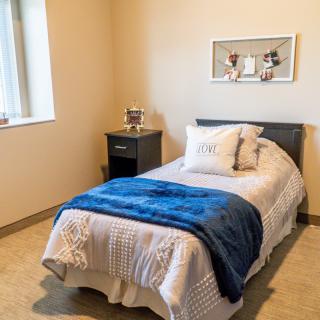 A cozy bedroom with a single bed, blue blanket, and decorative pillows next to a window and nightstand.