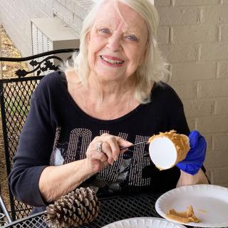 A smiling woman working on a craft project outdoors, using a pine cone.