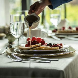 A hand pouring syrup over pancakes topped with fresh berries on a elegantly set table.