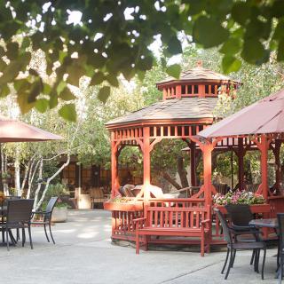 A serene outdoor garden setting featuring wooden gazebos and patio furniture under the shade of trees.