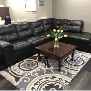A cozy living room featuring a black leather sectional sofa, a wooden coffee table with yellow tulips in a vase, and a patterned rug.