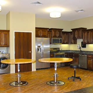 A modern kitchen with wood cabinets, stainless steel appliances, and round tables on a hardwood floor.