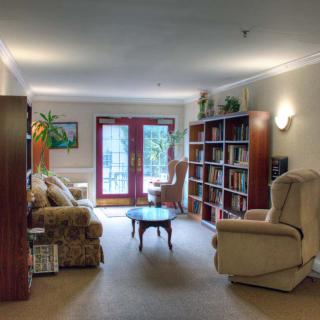 A cozy living room with armchairs, bookshelves, and a coffee table, illuminated by natural light from large glass doors.