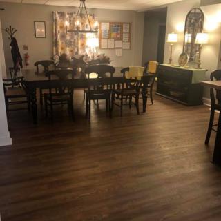 A cozy dining room with a wooden floor, featuring a large table surrounded by chairs, decorative lamps, and a chandelier.