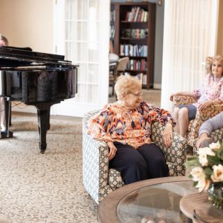 A cozy living room with seniors relaxing in armchairs, alongside a grand piano and bookshelves.