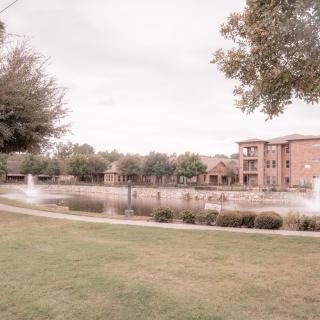 A serene landscape featuring a pond with fountains and an apartment building in the background.