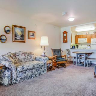 A cozy living room with a floral-patterned sofa, artwork on the walls, and a view of the kitchen area.