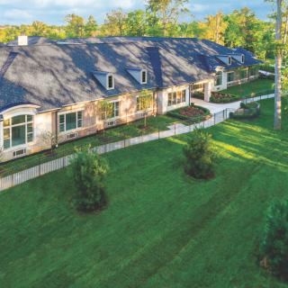 A long, elegant house with a sloped roof surrounded by a well-manicured lawn and trees.
