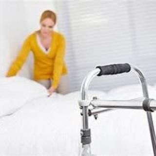 A woman making a bed with a walker in the foreground, suggesting a caregiving environment.