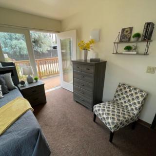 A cozy bedroom with a patterned chair, dresser, and decorative items on a shelf, featuring a large window with an outside view.