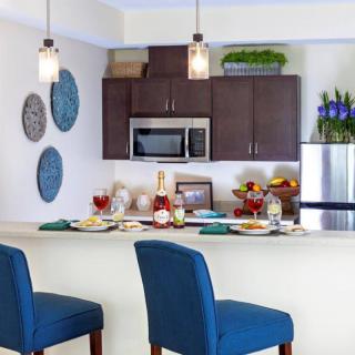 A modern kitchen interior featuring a countertop with blue bar stools, pendant lights, and a dining setup.