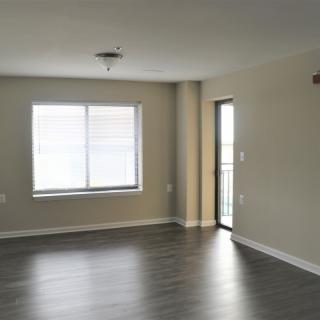An empty apartment room with hardwood flooring, a window, and a door leading outside.