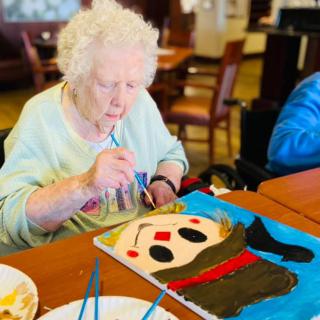 An elderly woman engaged in painting a colorful artwork at a table.
