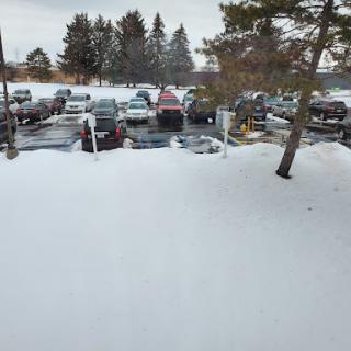 A snowy parking lot with several vehicles visible, surrounded by trees under a cloudy sky.