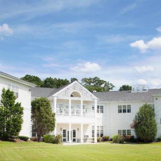 A residential building with a white exterior, surrounded by trees and a well-maintained lawn under a blue sky with clouds.