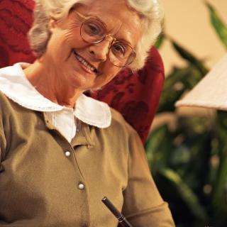 An elderly woman is sitting in a comfortable chair, smiling while holding a book, with a lamp and plants in the background.
