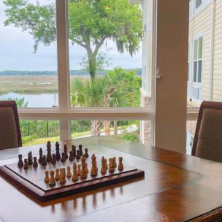 A chess board set up on a wooden table with a scenic view through large windows.
