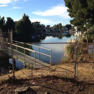 A view of a serene lake behind a chain-link fence, surrounded by trees and under a clear sky.