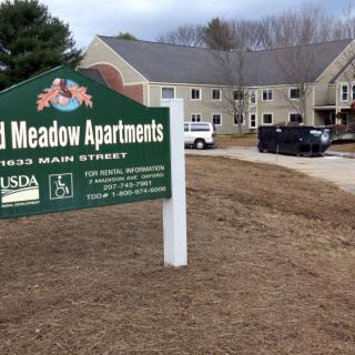 A sign for apartments in front of a residential building with parked vehicles.