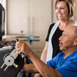 An elderly man exercises on a machine under the guidance of a trainer.