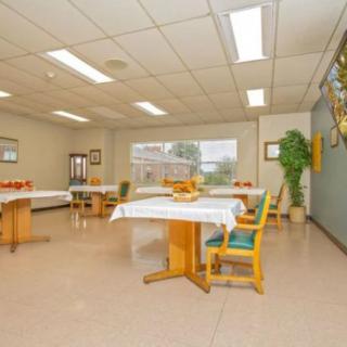 A spacious dining room with neatly arranged tables and chairs, well-lit by overhead fluorescent lights. Large windows allow natural light in, complemented by potted plants for a touch of greenery.
