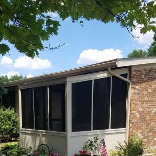 A house with a screened porch and a brick exterior, surrounded by trees and a garden under a blue sky.