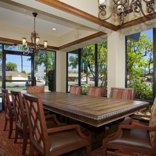 A spacious dining room with a large wooden table, surrounded by chairs and illuminated by chandeliers, featuring large windows that offer ample natural light.