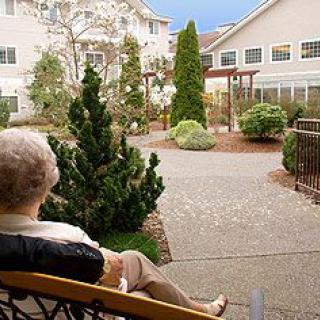 An elderly person sits on a bench, enjoying the serene view of a garden at a retirement home.